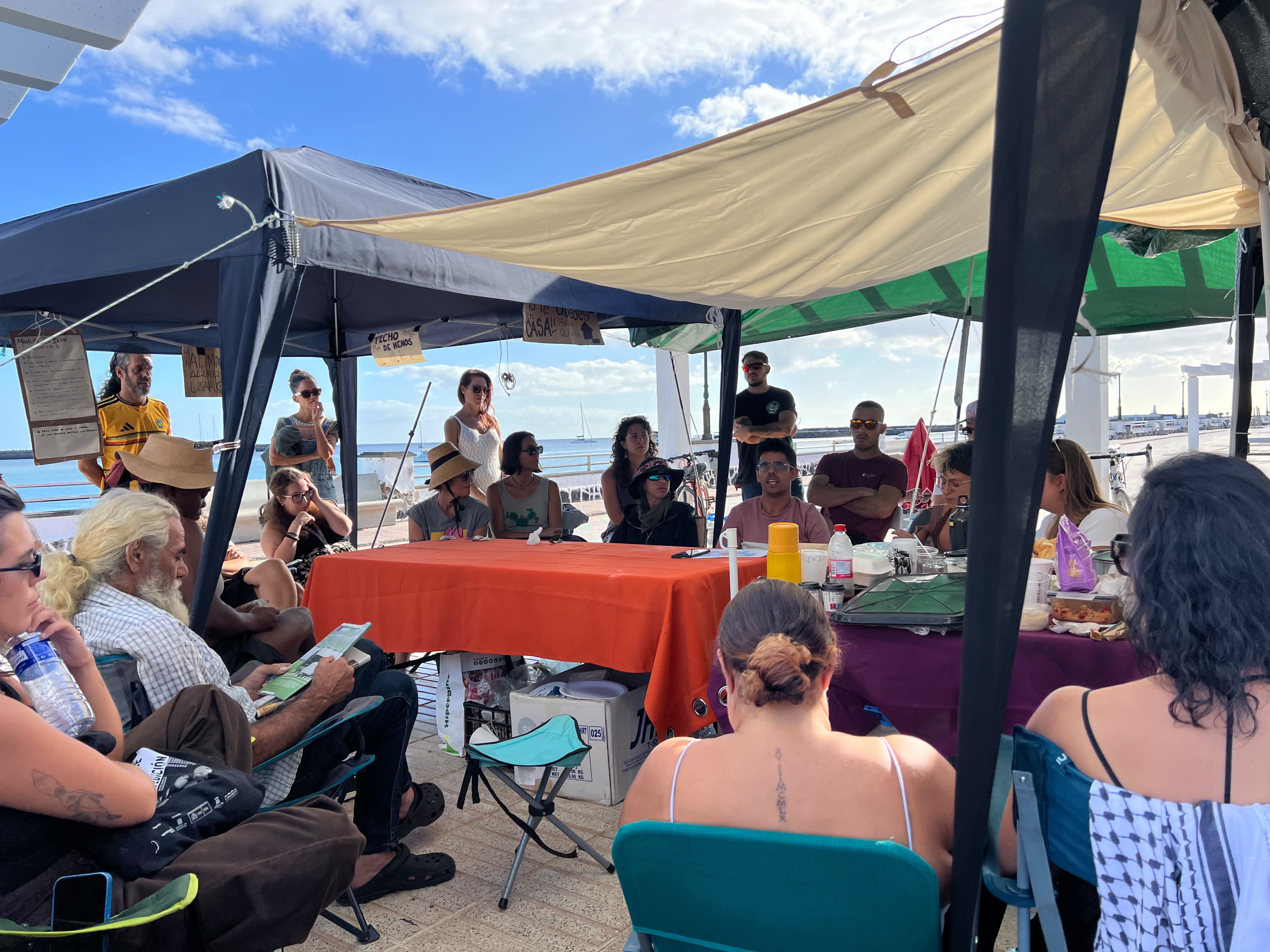 An assembly in the sun under the gazebos: people sitting in a circle around the table, with the sea behind