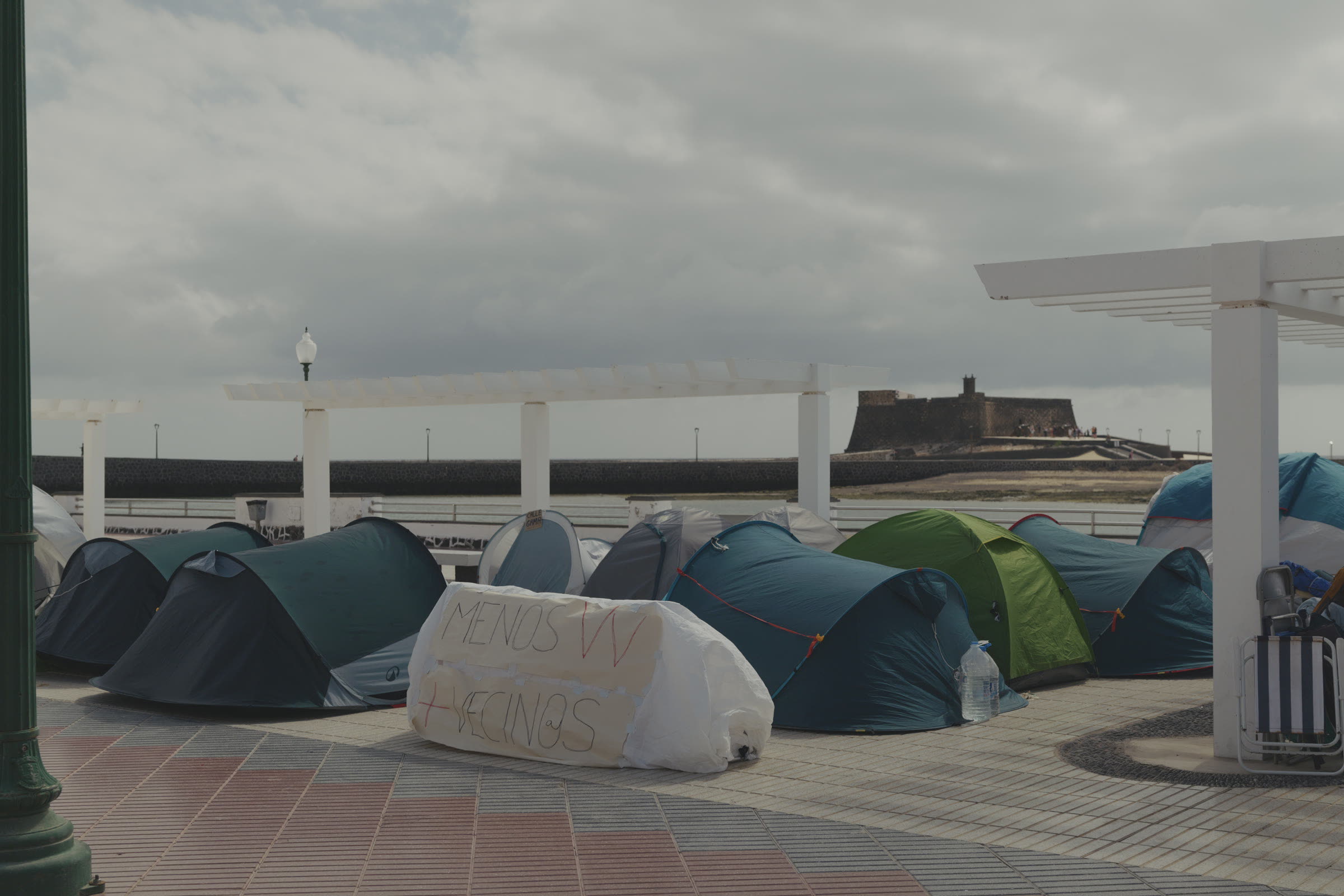 The camp's tents under the park pergola, one reading “Fewer holiday lets, more neighbours”, with the San Gabriel castle behind